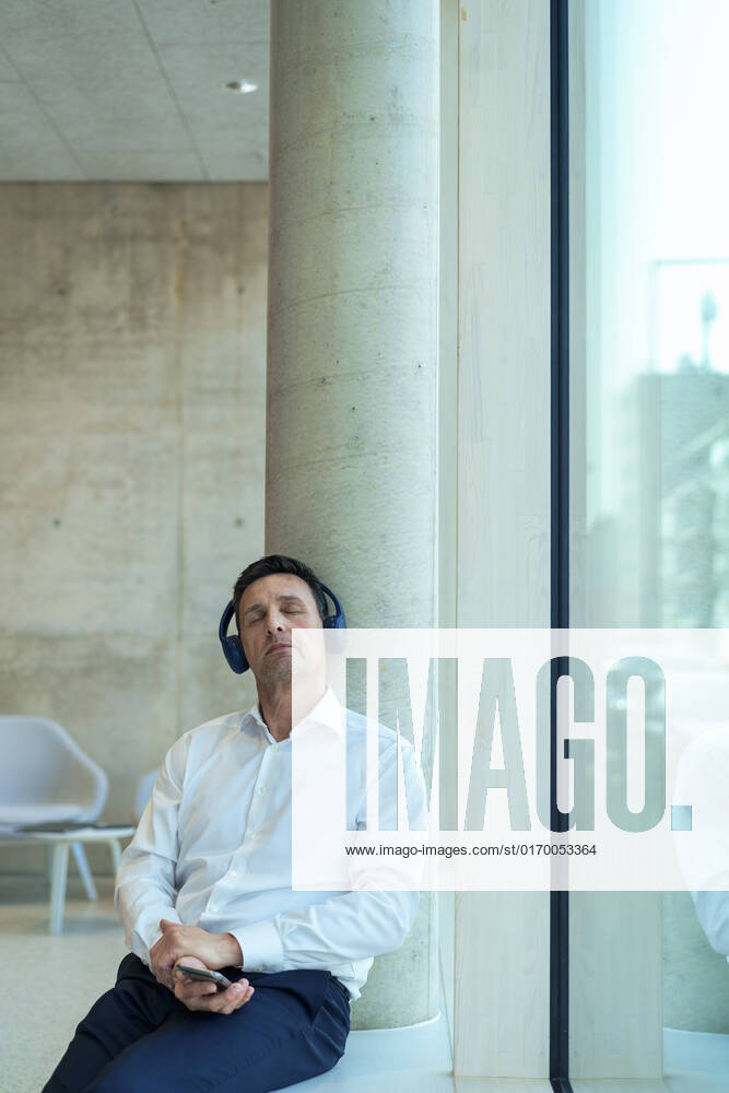 IMAGO Creative: 2022 Favoriten IMAGO Creative: Business - Progressive Geschäftsmodelle Businessman with headphones sitting by column near window at office model released, Symbolfoto property released, JOSEF12352
