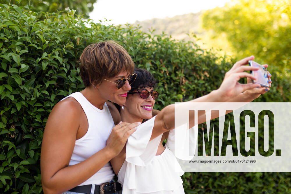 IMAGO Creative, Jahreszeit Sommer, Sommer in der Stadt Side view of stylish adult female friends standing near green shrubs in park and taking selfie on smartphone while spending summer weekend together PicuaEstudio_Lifestyle_18
