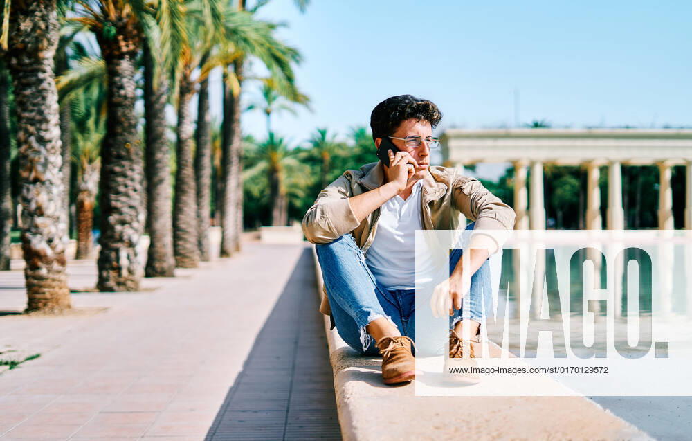 IMAGO Creative, Jahreszeit Sommer, Sommer in der Stadt Stylish pensive male having conversation of mobile phone while sitting near pond in tropical garden in city and looking away, Model released AdalbertoRodriguez_UniversityStudent_16