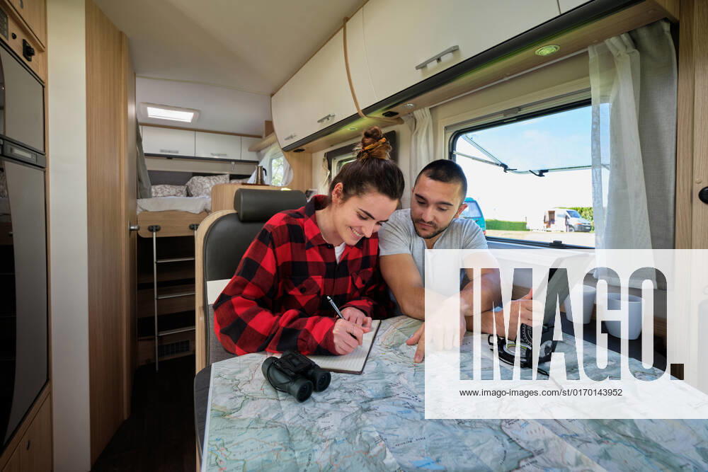 Cheerful couple of travelers planning route of trip while reading paper map and taking notes in notebook in caravan, Model released Pempki_CamperCouple_11