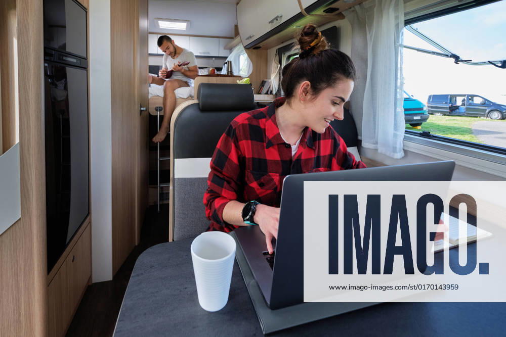 Content traveling female freelancer browsing laptop and taking notes in notebook while working remotely in camper on background of boyfriend, Model released Pempki_CamperCouple_14