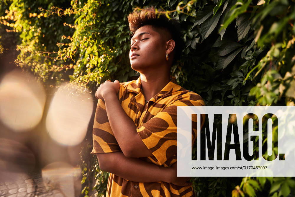 IMAGO Creative, Jahreszeit Sommer, Sommer in der Stadt Dreamy young Filipino non binary man wearing cool shirt with earring embracing himself with love standing with eyes closed in green garden ManuelRuiz_ID927_33969_031