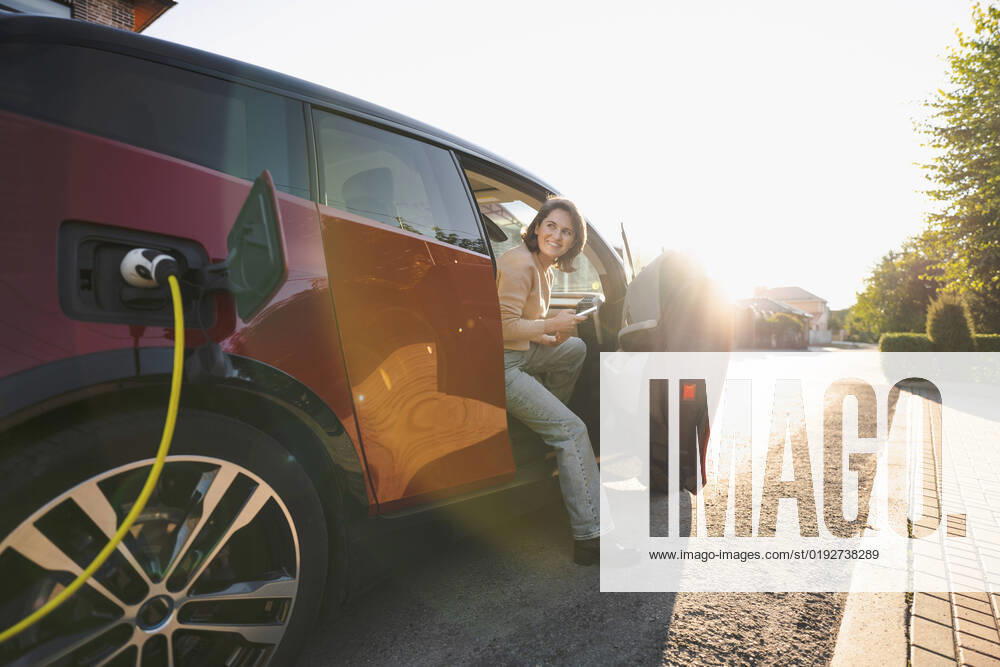 IMAGO Creative: Business, Innovative Technologie-Lösungen Happy woman with mobile phone sitting in electric car at vehicle charging station on sunny day model released, Symbolfoto, EKGF00152