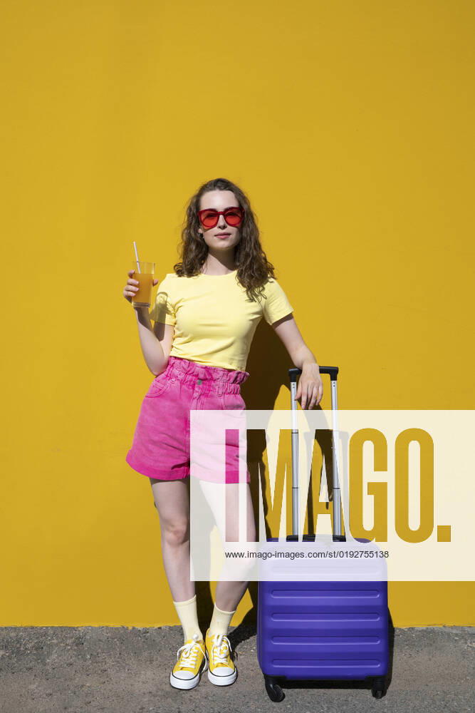 Woman wearing sunglasses standing with drink and luggage in front of yellow wall model released, Symbolfoto, VPIF07319