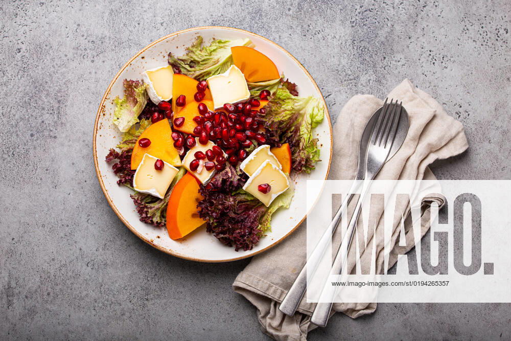 Healthy vegetarian persimmon salad with brie top view Top view flat lay persimmon salad with brie cheese, fresh salad leaves, pomegranate seeds on white plate and stone gray background, seasonal fruit salad as appetizer, vegetarian healthy food