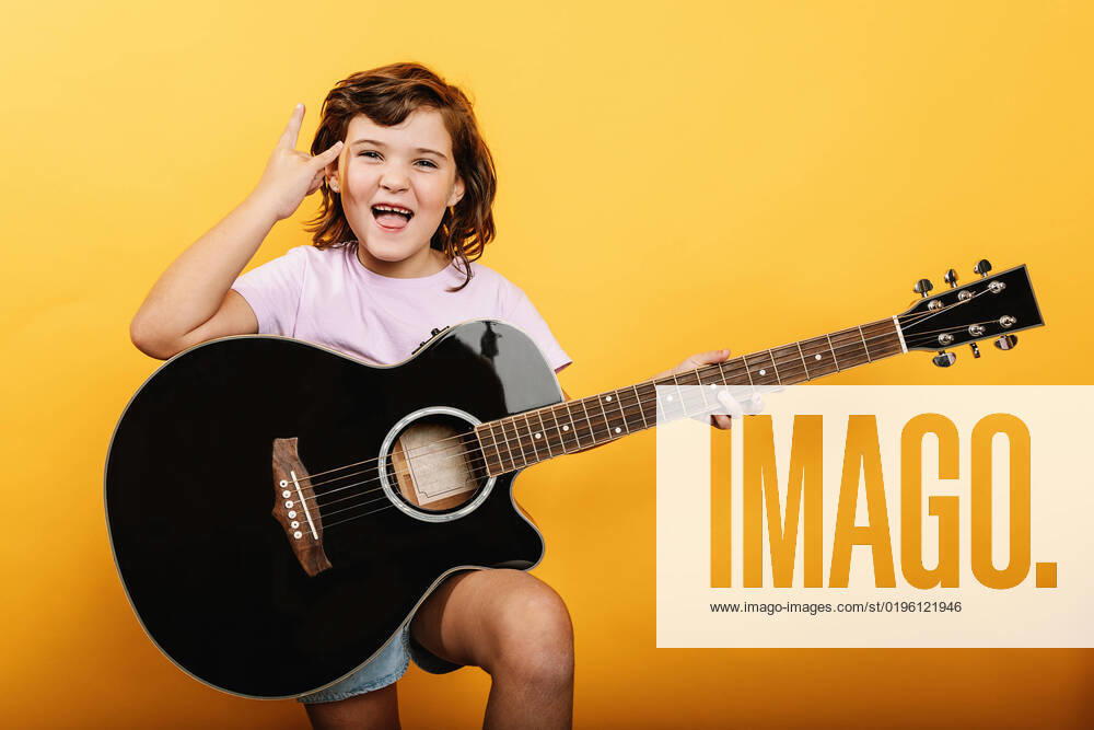 Happy kid in pink t shirt with mouth opened showing horn gesture while playing acoustic guitar against yellow background, Model released HodeiUnzueta_ID7769_234933_024