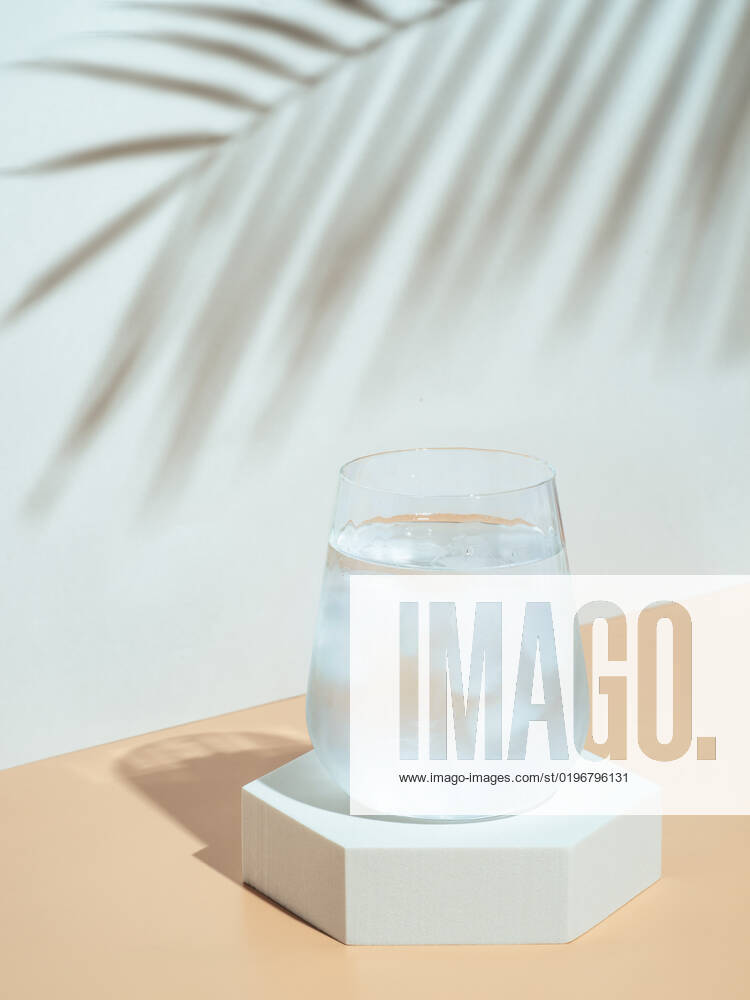 Modern still life with cold water glass Cold water with ice in tumbler glass on hexagon pedestal. Mockup for drink in fashion trendy style with tropical leaf shadow. Modern still life with glass. Copy space. Health, beverage, summer concept