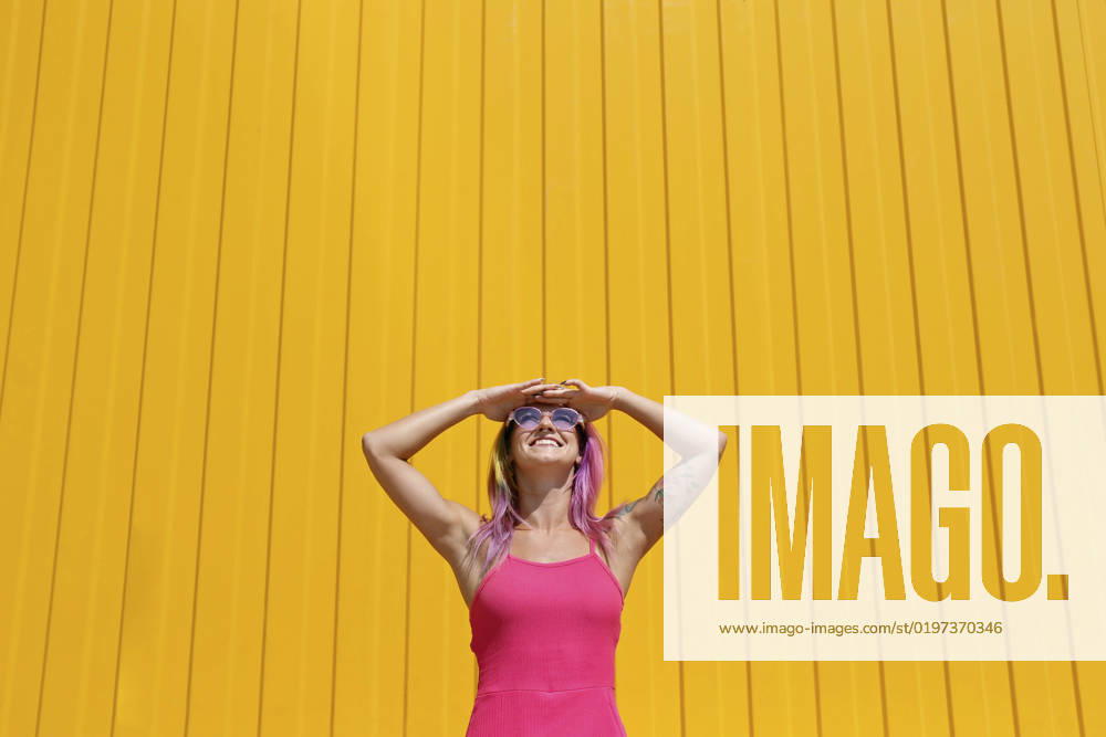 IMAGO Creative, Lifestyle, Barbiecore Happy woman wearing pink dress shielding eyes in front of yellow wall model released, Symbolfoto property released, SYEF00111