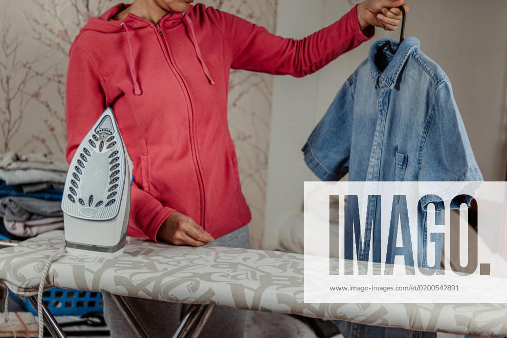 Crop unrecognizable female in casual outfit standing near ironing board with blue denim shirt on hanger while preparing clothes for work at home, Model released DavidGari_ID10740_325175_013