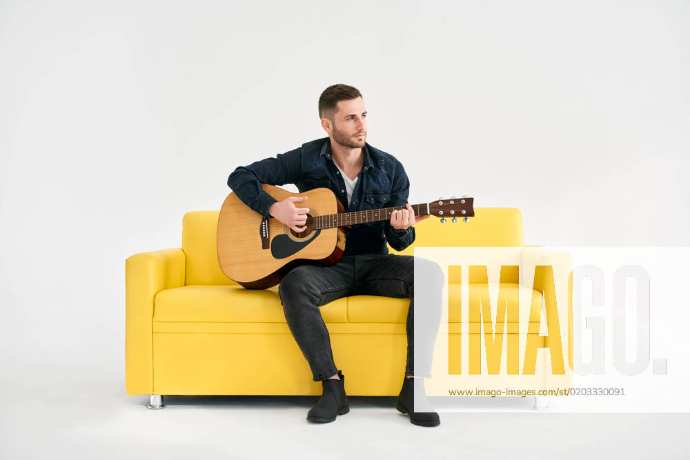 Handsome young man playing acoustic guitar whlile sitting on yellow sofa over white background Handsome young man playing acoustic guitar whlile sitting on yellow sofa over white background. Hobby, music concept