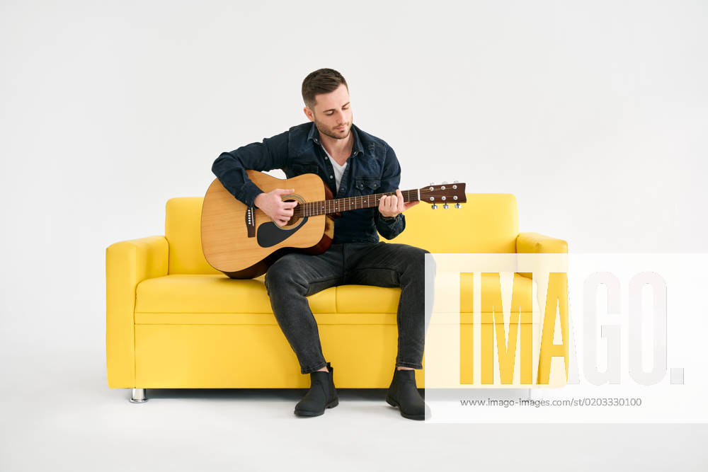 Handsome young man playing acoustic guitar whlile sitting on yellow sofa over white background Handsome young man playing acoustic guitar whlile sitting on yellow sofa over white background. Hobby, music concept