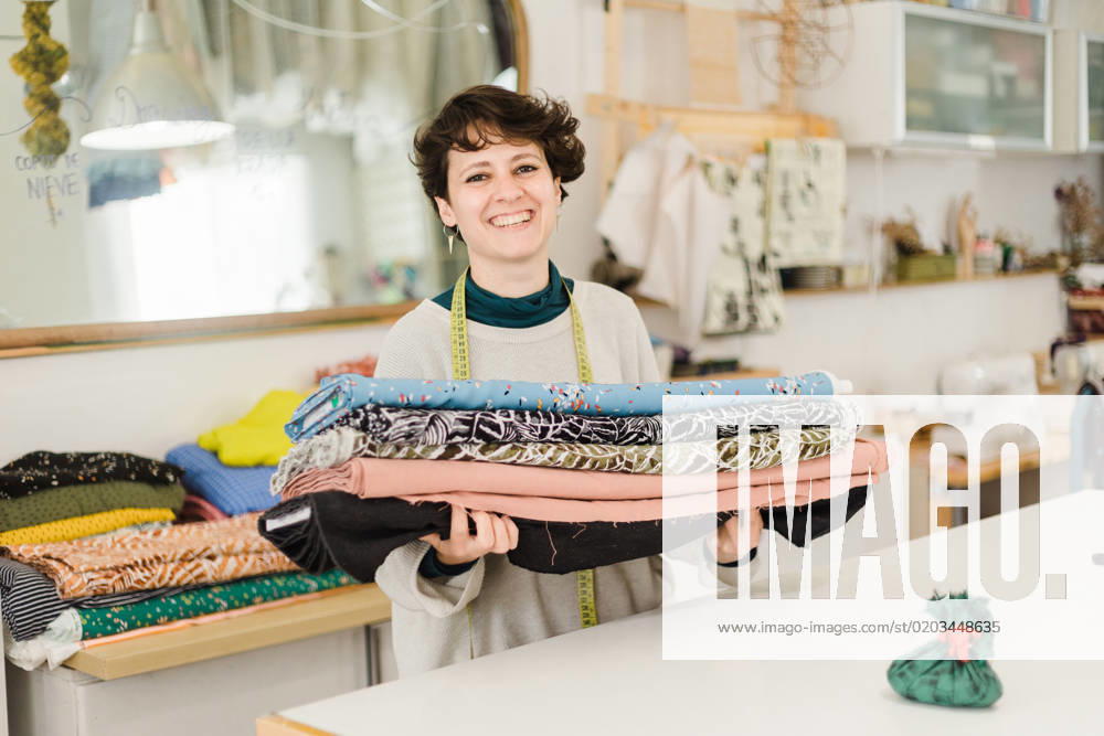 Cheerful female designer in casual clothes with measuring tape around neck looking at camera and smiling while standing with colorful textile in atelier, Model released, Property released AlbaCaro_ID12631_384029_058