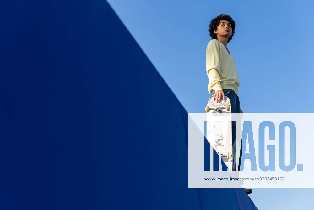 Low angle full body of thoughtful young Asian male skater in casual outfit standing on ramp with skateboard and looking away against blue cloudless sky, Model released DavidMunoz_ID12930_392769_042