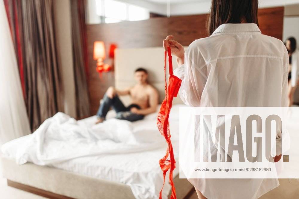 Sexy female person with red erotic bra in hand, man lies on big white bed. Intimate couple in bedroom, sex lovers. Sexy female with red bra in hand, itimate couple