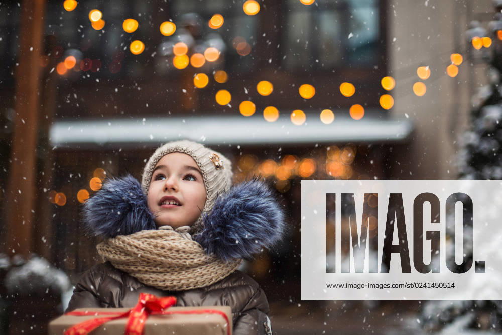 IMAGO Creative, Jahreszeit Winter in der Stadt Portrait of joyful girl with a gift box for Christmas on a city street in winter with snow on a festive market with decorations and fairy lights. Warm clothes, knitted hat, scarf and fur. New year Portrait of joyful girl with a gift box for Christmas on a city street in winter with snow on a festive market with decorations and fairy lights. Warm clothes, knitted hat, scarf and fur. New year ,model released, Symbolfoto