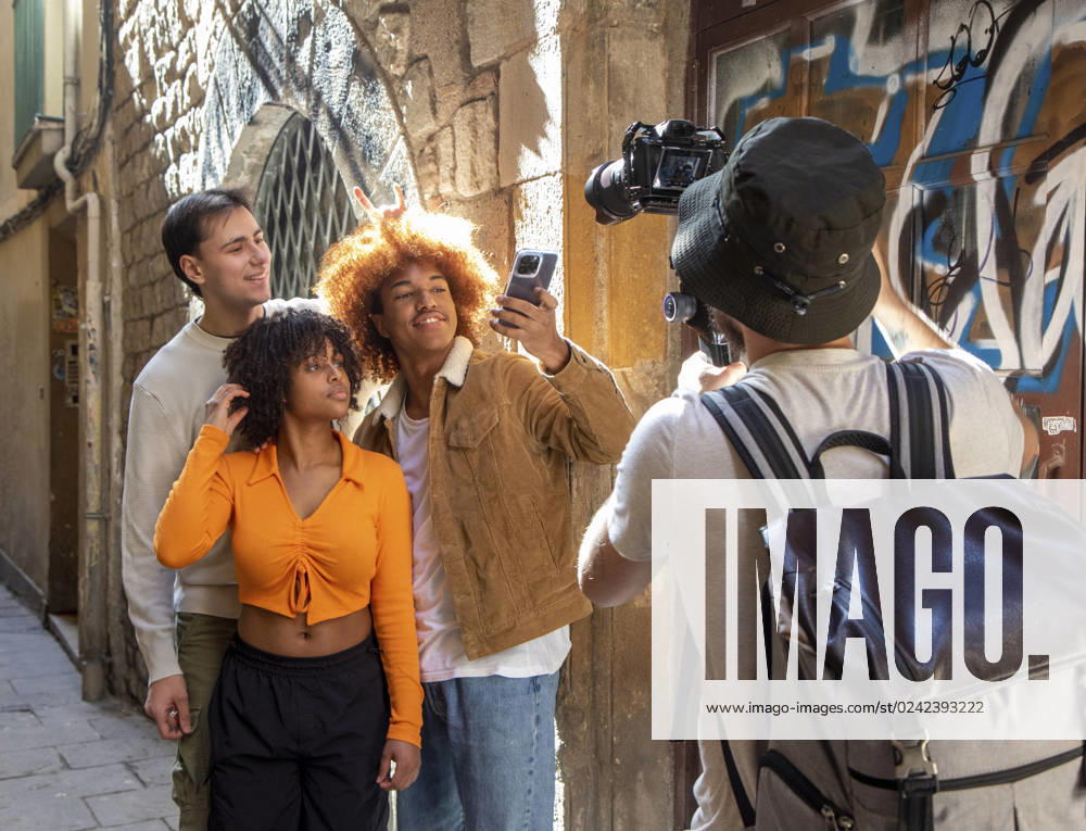 IMAGO Creative, Urban Lifestyle Back view of unrecognizable photographer taking picture of group of young multiethnic friends standing in street and looking at screen of mobile phone together while taking selfie, Model released LauraZamora_ID13393_408010_006