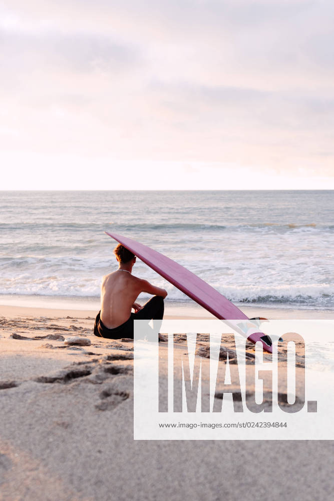 Back view of unrecognizable male surfer with naked torso and surfboard on head sitting on sandy coast and enjoying picturesque seascape during sunset, Model released NicoMortarini_ID13416_408708_014