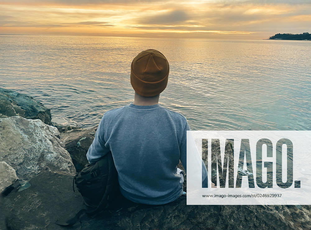 Young man in casual clothes sits on the rocks near the sea. Caucasian guy in a blue sweater and hat holds his jacket and looking at the calm sea. View from the back. Young man in casual clothes sits on the rocks near the sea. Caucasian guy in a blue sweater and hat holds his backpack and looking at the calm sea. View from the back. The concept of nature and peace. ,model released, Symbolfoto