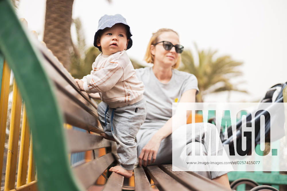IMAGO Creative, Urban Lifestyle Young mother with her cute infant baby boy child on bench on urban children s playground on warm summer day. Young mother with her cute infant baby boy child on bench on urban children s playground on warm summer day ,model released, Symbolfoto