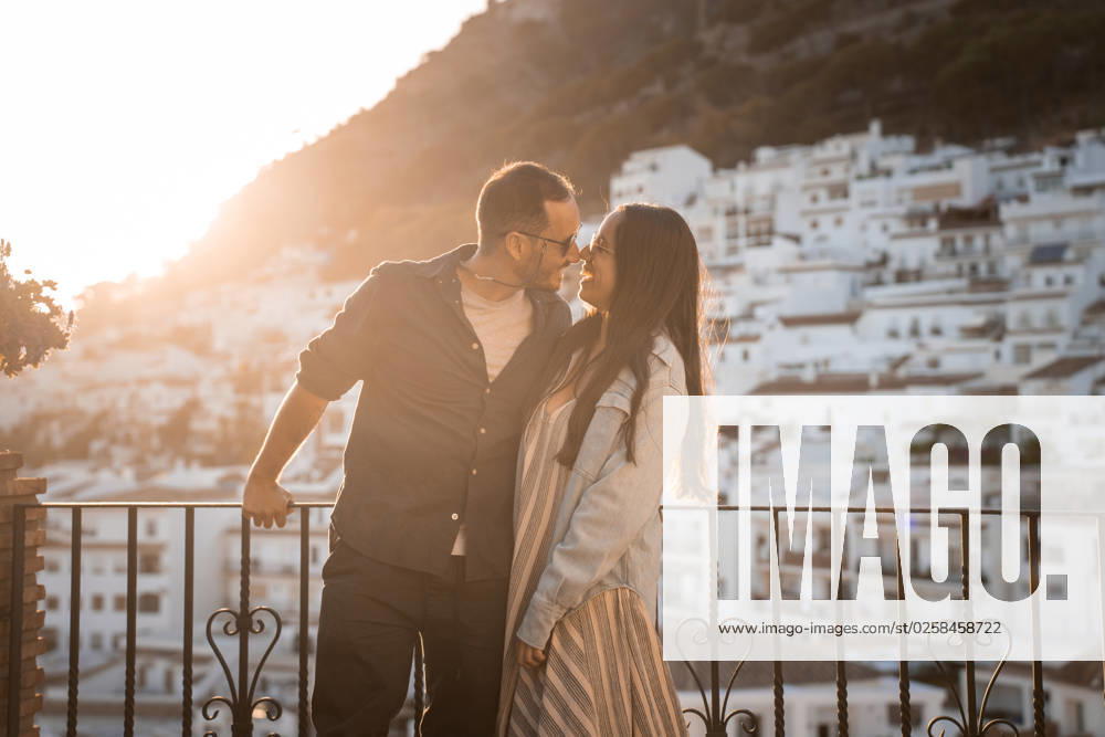 IMAGO Creative, Urban Lifestyle Loving couple standing close on balcony Happy couple in sunglasses hugging smiling on balcony with fence while looking at each other against city buildings and mountain during sunset time PabloMenendez_ID14105_425562_040