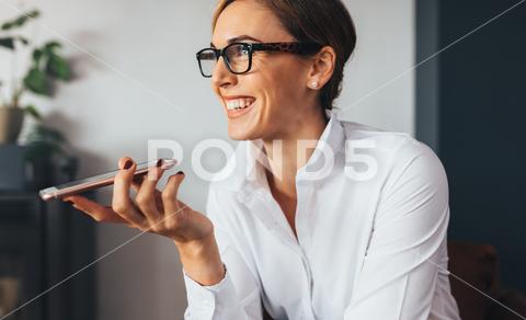 Closeup of smiling business woman talking over mobile phone on loudspeaker. Woman in formal attire holding a mobile phone close to her mouth and looking away. business, businesswoman, cell phone, close up, collar, entrepreneur, female, formal, g, happy, holding, indoors, lifestyle, looking away, loudspeaker, mobile phone, office, one person, people, phone, sitting, smart, smile, smiling, spectacles, talking, technology, using, white, wireless, worker, working, workplace, young woman