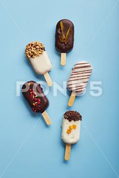 Mousse dessert with fruit flavors in the form of popsicle ice cream covered with white and dark chocolate on a blue background. ice cream, sweet, mousse, dessert, delicious, food, cream, popsicle, homemade, lollipop, cold, sugar, stick, pastry, party, pop, shape, vanilla, tasty, sprinkle, fruit, glac, inside, flake, variety, copy, cupcake, filling, space, stacked, assortment, bright, cheesecake, nobody, closeup, cookie, fresh, plate, cool, chocolate, decoration, cake, gastronomy, meal