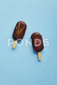 Mousse dessert with fruit flavors in the form of popsicle ice cream covered with dark chocolate on blue background. ice cream, sweet, mousse, dessert, delicious, food, cream, popsicle, homemade, lollipop, cold, sugar, stick, pastry, party, pop, shape, vanilla, tasty, sprinkle, fruit, glac, inside, flake, variety, copy, cupcake, filling, space, stacked, assortment, bright, cheesecake, nobody, closeup, cookie, fresh, plate, cool, chocolate, decoration, cake, gastronomy, meal