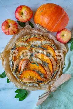 A homemade pumpkin quiche on the table with a pumpkin and apples apples,autumn,background,baked,cake,celebration,cheesecake,cinnamon,cooked,cooking,cuisine,delicious,dinner,food,fresh,gastronomy,gourmet,grilled,halloween,homemade,lunch,meal,plate,pumpkin,quiche,table,tasty,top view,traditional,vegetable,vegetarian,vertical