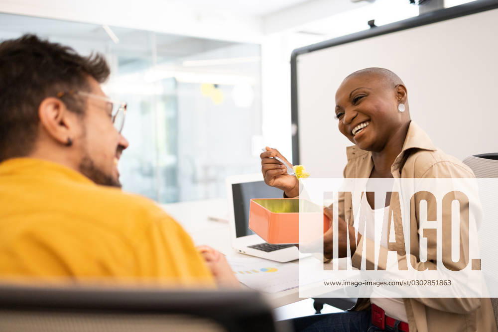 Happy black woman with a plate and spoon eating while sitting with a smiling man looking at each other under the office lights Model Released DavidMunoz_ID16062_488872_078