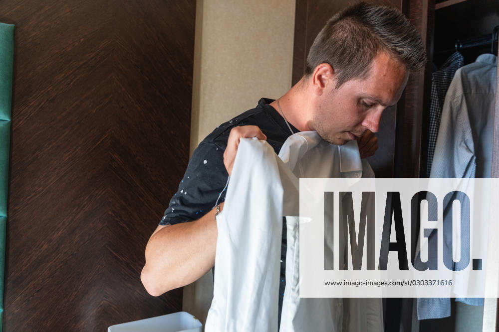 Young man holds a white shirt in front of a closet and looks at it. Trying on clothes