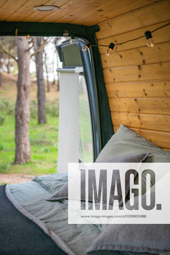 Tourist van with cozy bed pillows blanket near wooden planks wall ornament lights opened door with sun shade against blurred Andalusia trees Model Released LuisPina_ID16420_499229_019
