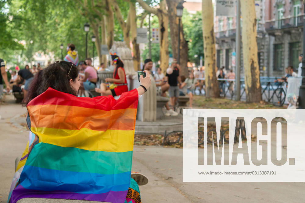 IMAGO Creative: Sommer, Sommertage im Stadtpark MR_ID17208_15471_JimenaPardo.jpg Side view of woman wraps herself with a large and colorful LGBTQ pride flag while taking selfie with her smartphone in a park with people in the background Model Released NataliaPenado_ID17208_523683_015