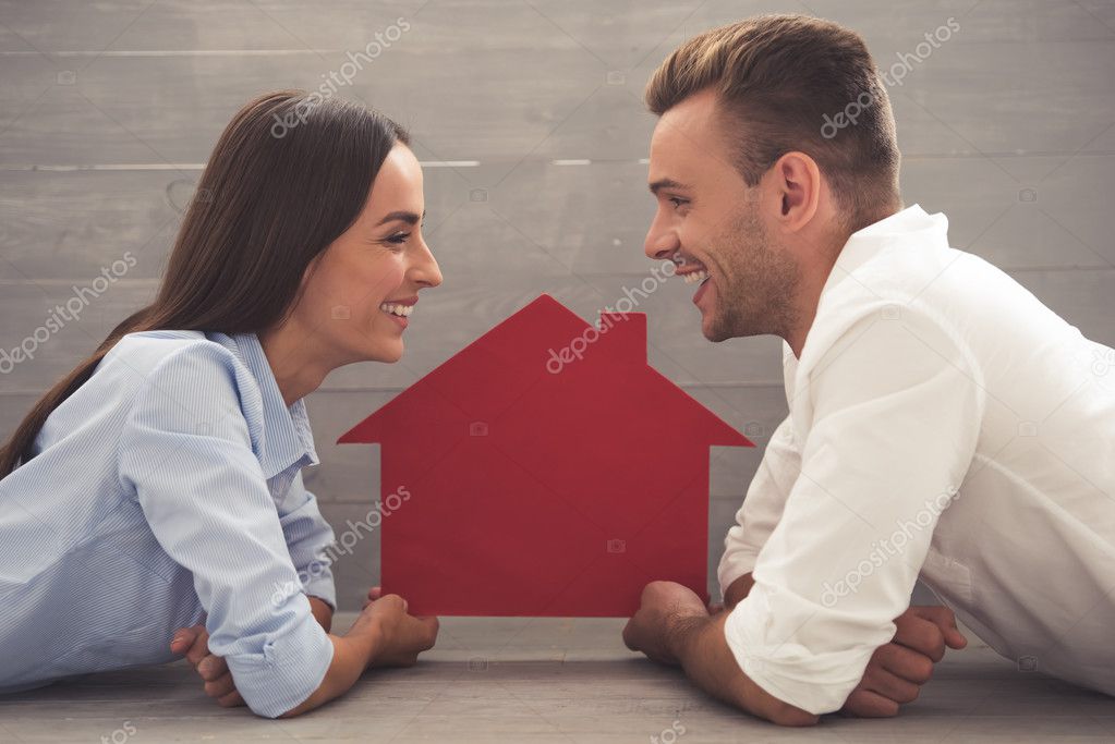 Side view of beautiful young couple in classic shirts holding a paper house, looking at each other and smiling while lying on the floor at home model released