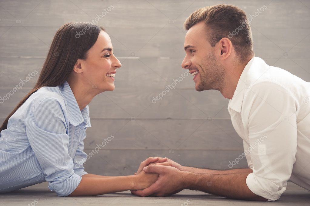 Side view of beautiful young couple in classic shirts holding hands, looking at each other and smiling while lying on the floor at home model released