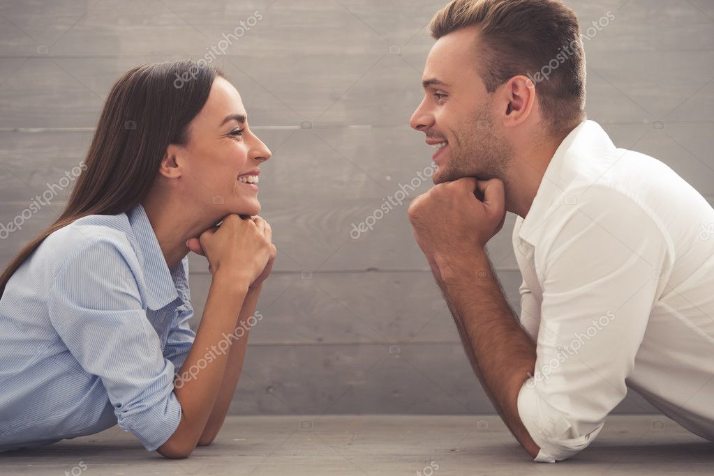 Side view of beautiful young couple in classic shirts leaning on their hands, looking at each other and smiling while lying on the floor at home model released