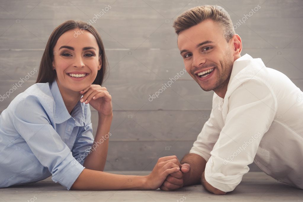 Side view of beautiful young couple in classic shirts holding hands, looking at camera and smiling while lying on the floor at home model released