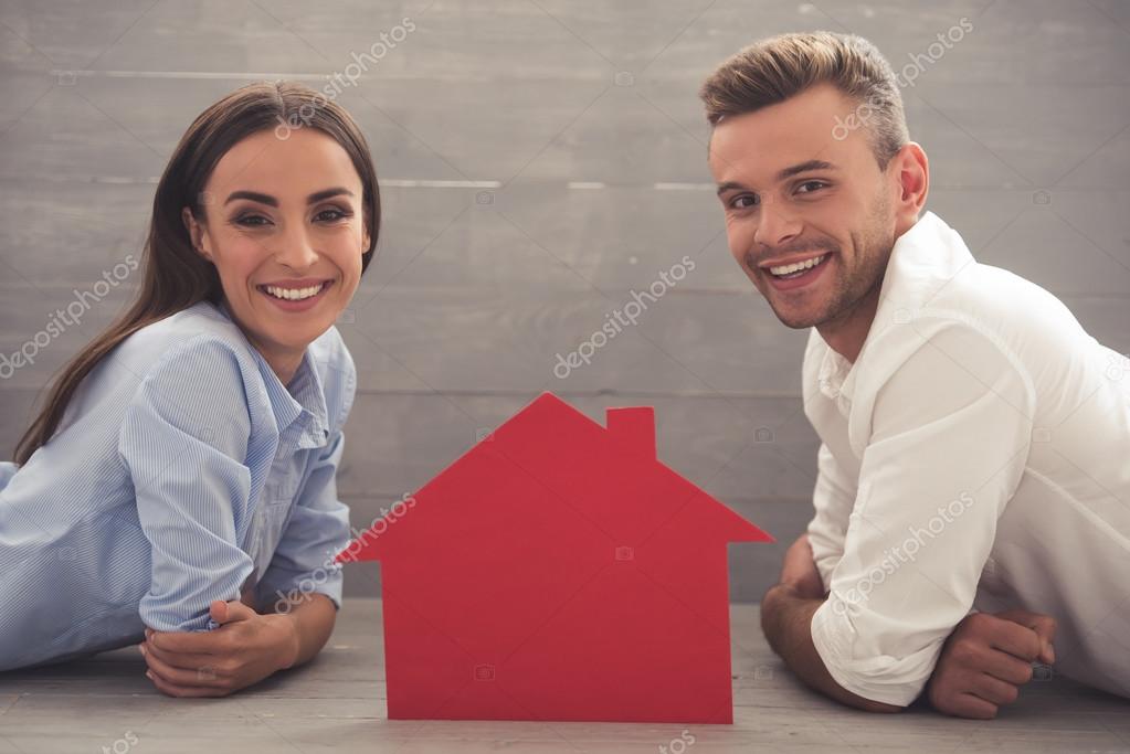 Side view of beautiful young couple in classic shirts looking at camera and smiling while lying on the floor at home. A paper house between them model released