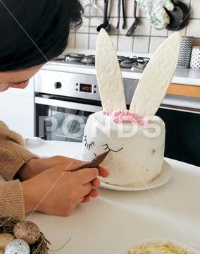 Person baking and decorating cake shaped as a bunny. kitchen, home, bake, baking, sweet food, cake, desert, preparing food, piping, piping bag, white chocolate, easter cake, close-up, one person, unrecognizable person, ingredients