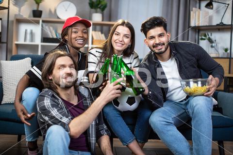 Group of multiracial friends toasting with beer while sitting on cozy sofa during european football match. Young men and women enjoying soccer game at home. {friends,diverse,cheering,excitement,gesturing,television,four,match,soccer,watch,fan,football,european,couch,support,community,enjoyment,indian,team,emotion,competition,entertainment,together,african,room,game,championship,group,smiling,happiness,sitting,multiracial,sport,drinking,fun,beer,man,home,woman,young,caucasian,leisure,eating,multicultural}