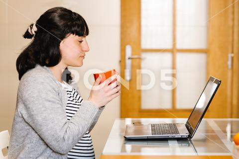 Side view of young ethnic pregnant woman with dark hair in casual clothes drinking hot coffee and watching interesting video on laptop while sitting at table in kitchen {{woman,watch,laptop,pregnant,drink,coffee,kitchen,expect,breakfast,video,internet,beverage,female,young,ethnic,hispanic,brunette,motherhood,pregnancy,netbook,home,social media,digital,maternal,free time,lady,online,casual,prenatal,await,hot drink,apartment,anticipate,connection,read,freelance,self employed,telework,lifestyle,indoors,side view,looking at screen,blurred background,horizontal}}