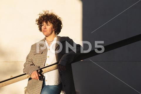 Confident female in trendy outfit looking into distance while leaning on railing near white wall with shadow on sunny street {woman,style,street,railing,wall,fashion,appearance,outfit,feminine,city,garment,shadow,trendy,sunlight,confident,building,female,sunshine,lady,gorgeous,attractive,apparel,pensive,charming,attire,town,light,wear,elegant,construction,district,bright,wistful,dreamy,thoughtful,sunbeam,ponder,design,shade,summer,charm,horizontal,outside}