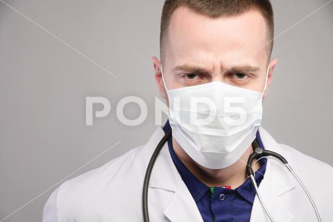 The stern look of a young doctor in a protective mask looks sullenly into the camera. studio portrait. isolated, adult, serious, portrait, confident, caucasian, angry, glasses, man, work, white, tie, corporate, executive, eyebrows, imperious, leader, mature, power, proprietor, red, spectacles, supervisor, leadership, mentor, stern, brown, business, boss, suit, doctor, medical, hospital, face, looking