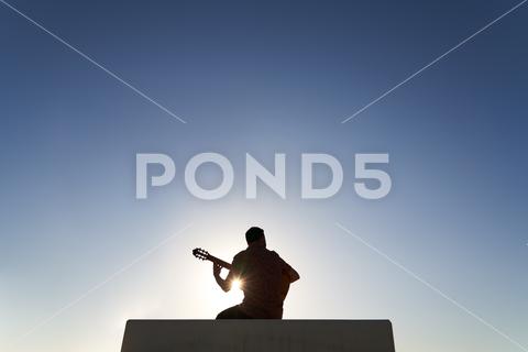 Back view silhouette of unrecognizable bearded musician playing song on acoustic guitar while sitting on border against blue cloudless sky on sunny day in city man, guitarist, guitar, silhouette, play, musician, music, song, instrument, hobby, acoustic, city, practice, talent, male, skill, sunset, perform, melody, border, cloudless, beard, town, sunlight, player, blue sky, sound, chord, barrier, summer, string, entertain, creative, sundown, instrumental, artist, sunshine, summertime, enjoy, outdoors, spend time, shadow, live, lifestyle, anonymous, unrecognizable, faceless, back view, spare time