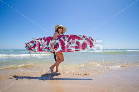 A Hispanic woman wearing swimwear while holding a surfboard on the beach attractive, beach, coast, female, girl, happy, hispanic, holiday, leisure, lifestyle, ocean, people, person, pretty, sand, sea, summer, surf, swim, vacation, voard, water, wear, woman, young