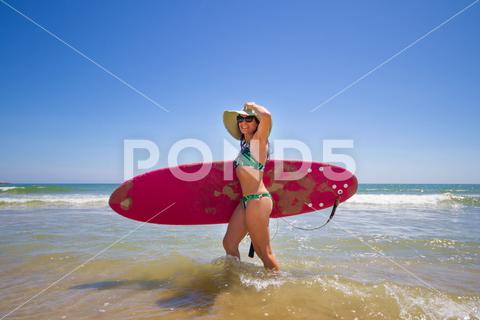 A Hispanic woman wearing swimwear while holding a surfboard on the beach attractive, beach, coast, female, girl, happy, hispanic, holiday, leisure, lifestyle, ocean, people, person, pretty, sand, sea, summer, surf, swim, vacation, voard, water, wear, woman, young