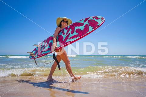 A Hispanic woman wearing swimwear while holding a surfboard on the beach attractive, beach, coast, female, girl, happy, hispanic, holiday, leisure, lifestyle, ocean, people, person, pretty, sand, sea, summer, surf, swim, vacation, voard, water, wear, woman, young