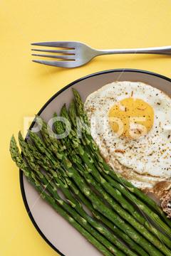From above breakfast dish of fried egg with green asparagus server on plate on yellow background egg, asparagus, breakfast, lunch, healthy, food, meal, top view, fresh, diet, fried, delicious, vegetable, cooking, appetizer, dinner, green, cuisine, dish, recipe, nutrition, organic, cooked, tasty, natural, plate, snack, menu, gourmet, table, protein, fat, pepper, spice, copy space, yellow background, from above, overhead, brunch, fork