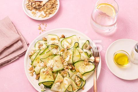 From above fresh salad of thin sliced ripe zucchini, feta cheese and pine nuts in white plate on table pink background zucchini, lemon, feta cheese, summer food, summer, vitamin, mix, organic, ingredient, mixed, oil, olive, vegetarian, homemade, zoodles, mozzarella, pine nuts, fork, healthy, gourmet, water glass, mediterranean, fresh, food, green, diet, table, recipe, meal, salad, herb, raw, culinary, nutrition, cuisine, tasty, gastronomy, vegetable, dish, lunch, traditional, dinner, delicious, noodles, nature, kitchen, pink background, from above