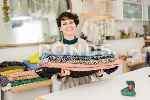 Cheerful female designer in casual clothes with measuring tape around neck looking at camera and smiling while standing with colorful textile in atelier {{{woman,designer,dressmaker,atelier,fabric,workshop,measure,textile,smile,studio,craft,tape,creative,cloth,seamstress,cheerful,professional,work,tailor,design,small business,style,female,handmade,handicraft,positive,fashion,workplace,profession,inspiration,happy,occupation,craftswoman,master,content,manufacture,skill,create,workspace,sew,material,project,talent,colorful,stand,parlor,workroom,looking at camera,blurred background}}}