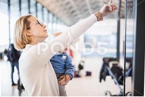 Mother traveling with child, holding his infant baby boy at airport terminal, checking flight schedule, waiting to board a plane. Travel with kids concept airport, travel, journey, infant, flight, mom, mother, terminal, woman, tourist, passenger, departure, looking, tourism, traveler, holiday, people, vacation, family, screen, gate, schedule, transport, view, transportation, flying, love, holding, electronic, business, tourists, domestic, passengers, airline, waiting, stroller, boy, luggage, kid, toddler, real people, station, train, commute, hold, parent, metro